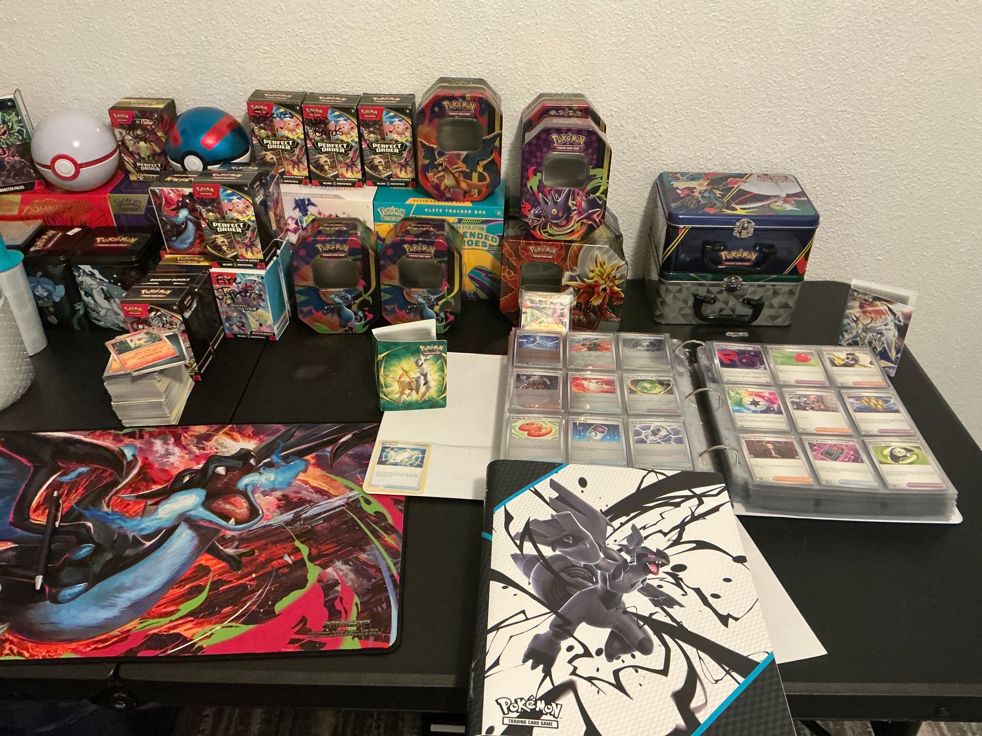Pokémon TCG tins, booster bundles, an Elite Trainer Box and binders of sleeved cards laid out on a table, with a Charizard playmat in front